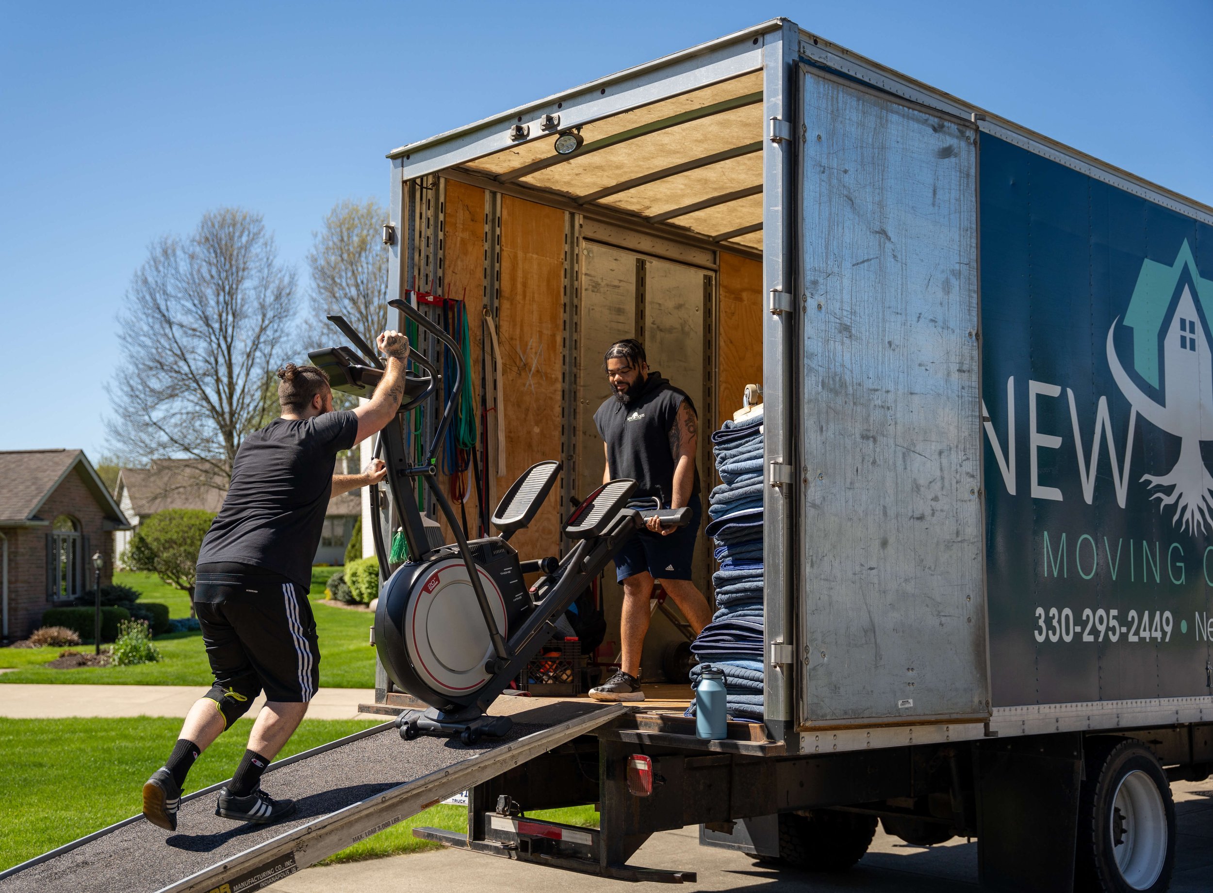 Two movers are loading a piece of exercise equipment, specifically an elliptical machine, into the back of a moving truck. One mover is pushing the equipment up a ramp, while the other is inside the truck, ready to guide it in. The truck has the branding 'New Roots Moving Company.' on the side, with the company's contact information visible. The scene takes place in a suburban neighborhood on a clear day.