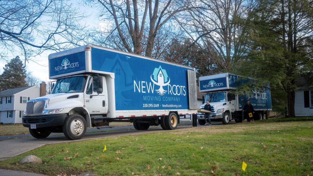 Two moving trucks from New Roots Moving Company are parked on a residential street, ready for a moving job. Three team members are seen in the background, standing near the trucks, prepared for the moving process.